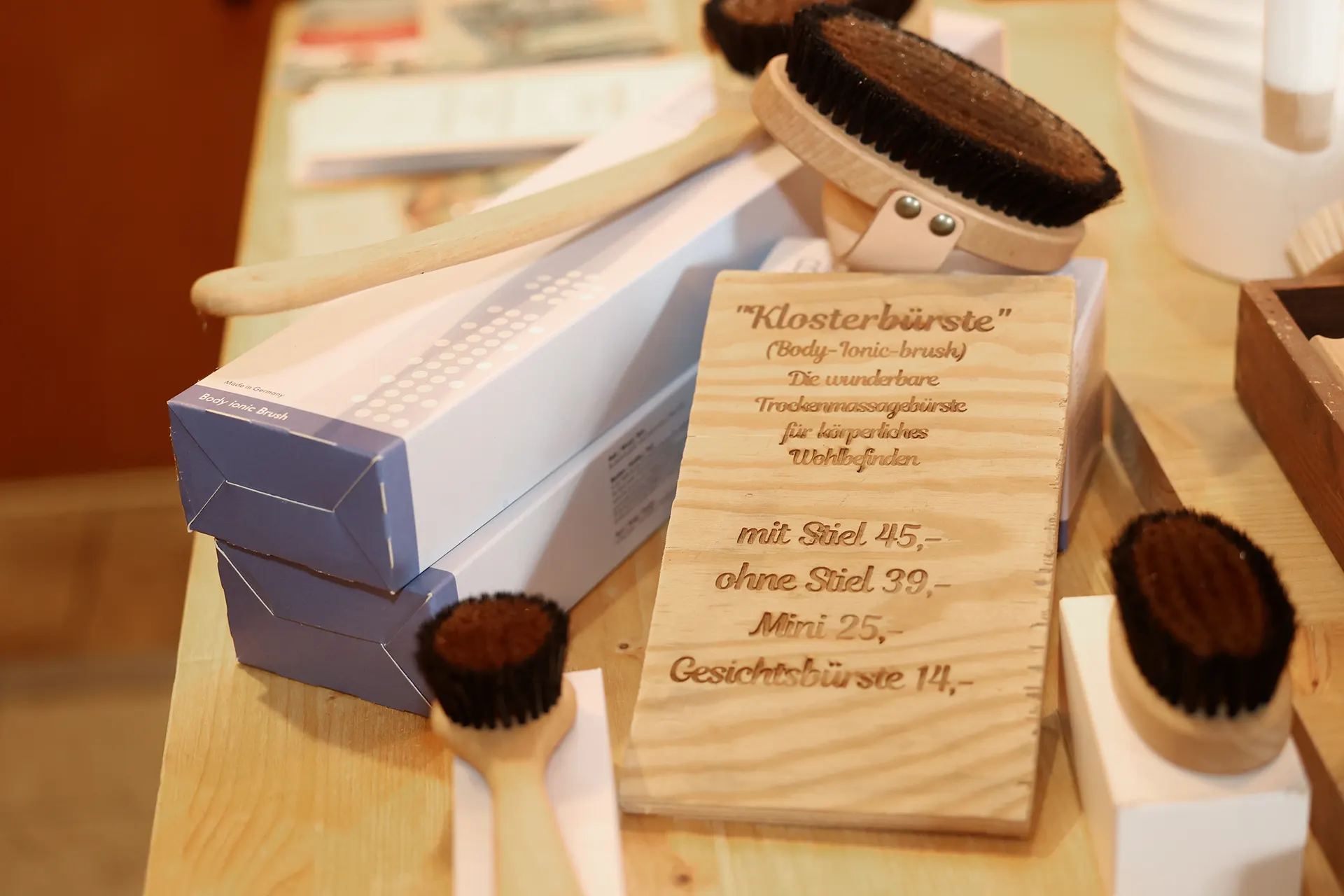 Klosterbürste (Body-Ionic-Brush) in der Ladenauslage mit graviertem Holzschild „Die wunderbare Trockenmassagebürste“, erhältlich mit Stiel, ohne Stiel, als Mini und als Gesichtsbürste